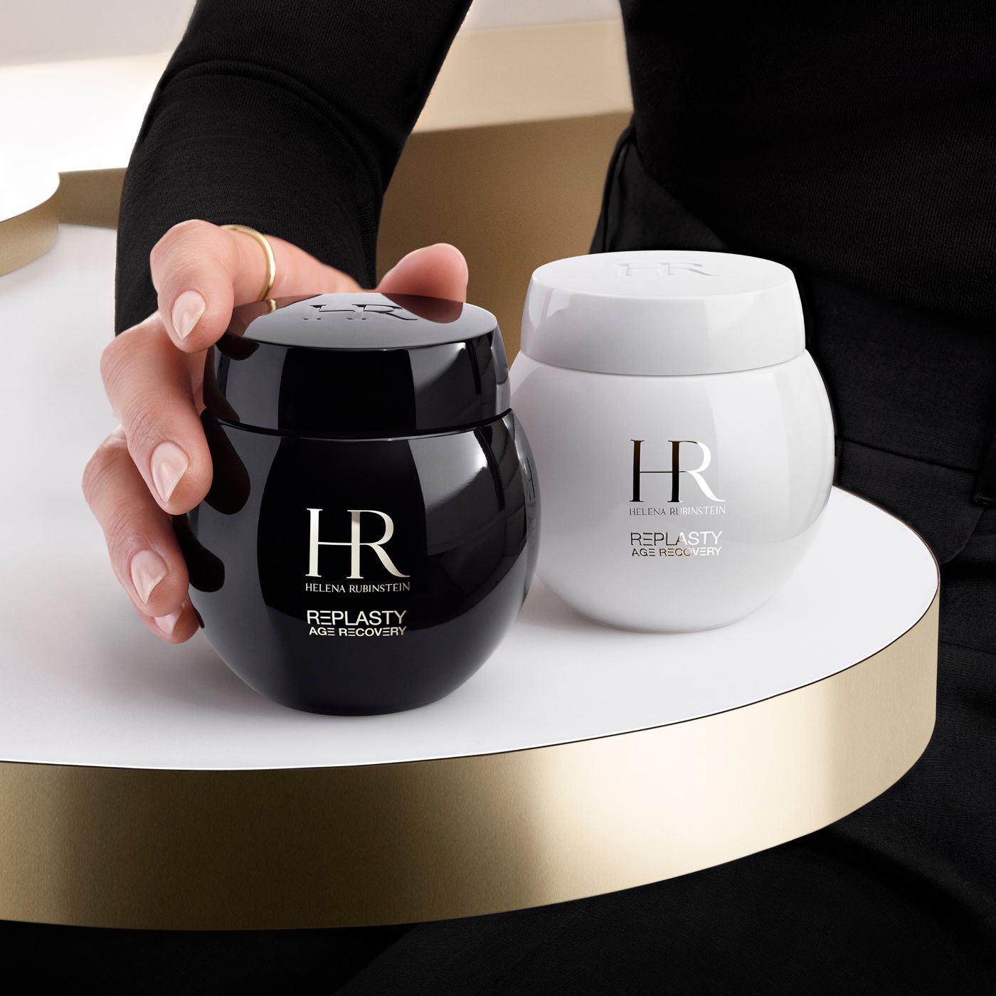 Eine Hand ergreift einen schwarzen Tiegel der Helena Rubinstein Re-Plasty Age Recovery Creme, ein passender weißer Tiegel daneben, auf einer goldenen Plattform.