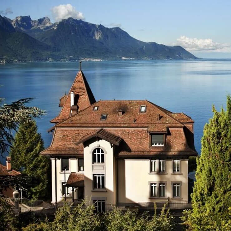 Ein großes Haus im traditionellen Stil mit rotem Ziegeldach und hellen Wanden, gelegen an einem großen tiefblauen See, umgeben von Bergen. Der Himmel ist klar.