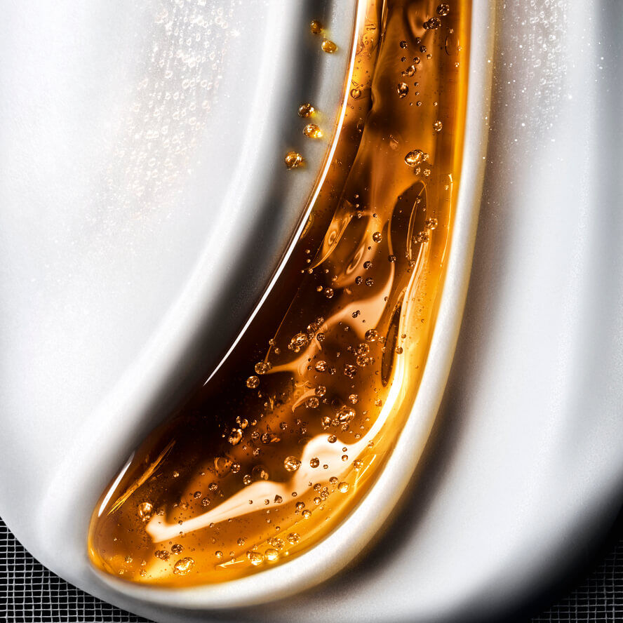 Nahaufnahme einer flussigen Kosmetiktextur, die schimmernde goldene Gelwellen mit Blaschen auf cremig-weißem Hintergrund mischt, was eine reichhaltige Pflege suggeriert.