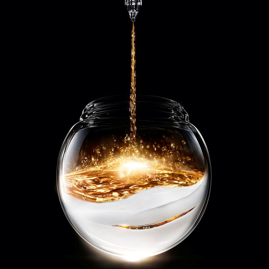 Funkelnde goldene Flussigkeit gießt in ein kugelformiges Glasgefaß, das auf einer texturierten weißen Basis ruht und einen leuchtenden Kontrast auf schwarzem Hintergrund erzeugt.