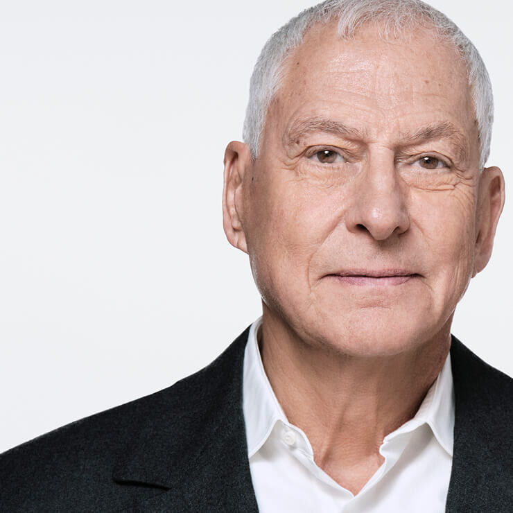 Nahaufnahme eines alteren Mannes mit grauem Haar, weißem Hemd und dunklem Sakko, leicht zur Seite blickend. Sein Ausdruck ist neutral und sein Gesicht ist beleuchtet.