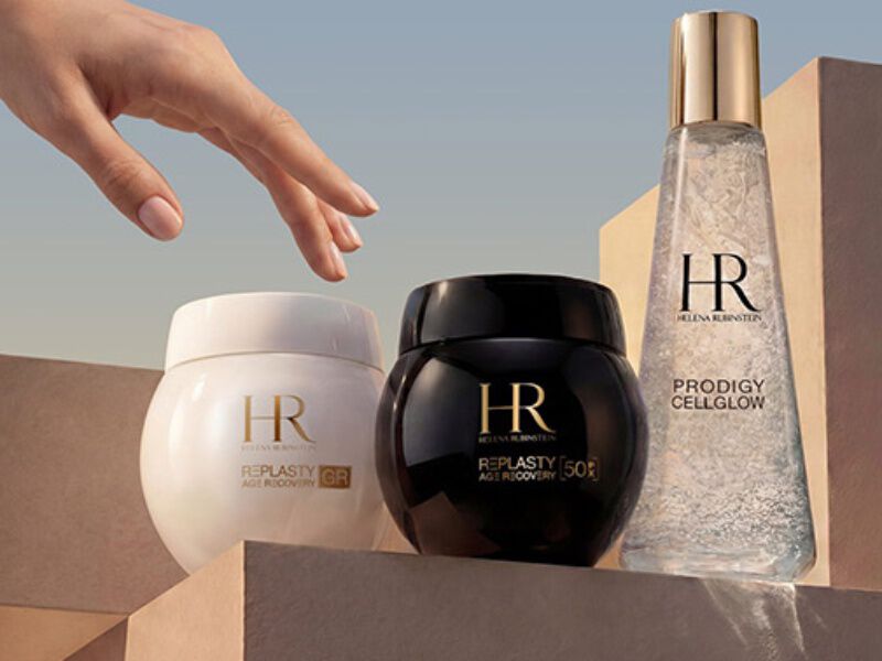 Eine Hand streckt sich nach drei Produkten von Helena Rubinstein aus, die sich durch ein elegantes Design und sanfte Beleuchtung auszeichnen.