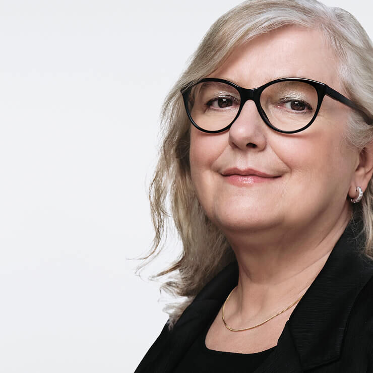 Nahaufnahme einer mittelalten Frau mit hellgrauem Haar, die eine schwarze Brille und eine dezente Goldkette tragt und leicht lachelt.