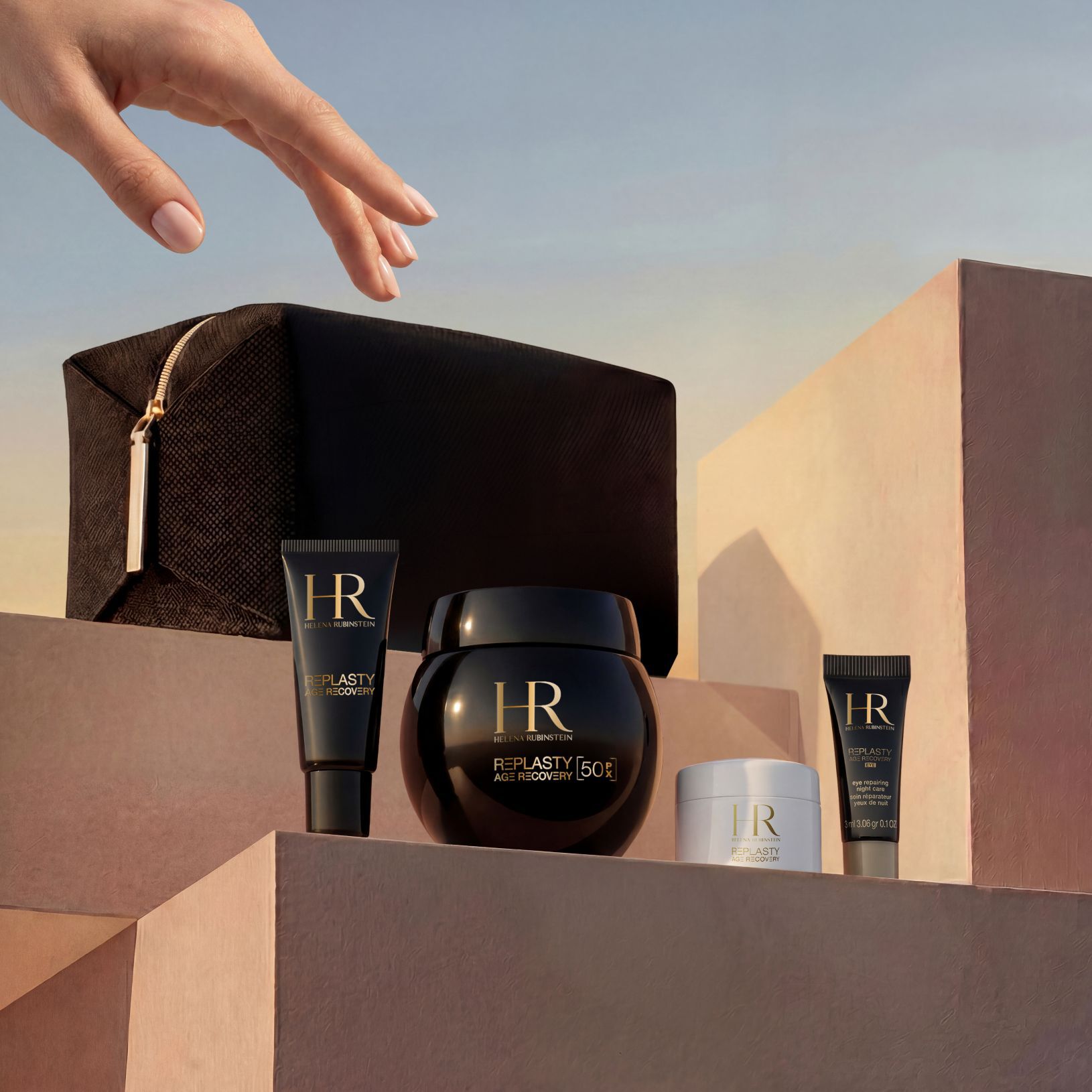 Eine elegante Hand nahert sich einem Helena Rubinstein Geschenkset, das auf sandfarbenen Steinblocken vor einem Sonnenuntergangshimmel prasentiert wird und ein schwarzes Reisetaschchen in limitierter Auflage sowie die Replasty Age Recovery Reihe umfasst: Eye Repairing Night Care, Day Cream, Night 50PX und Body Repairing Care.​