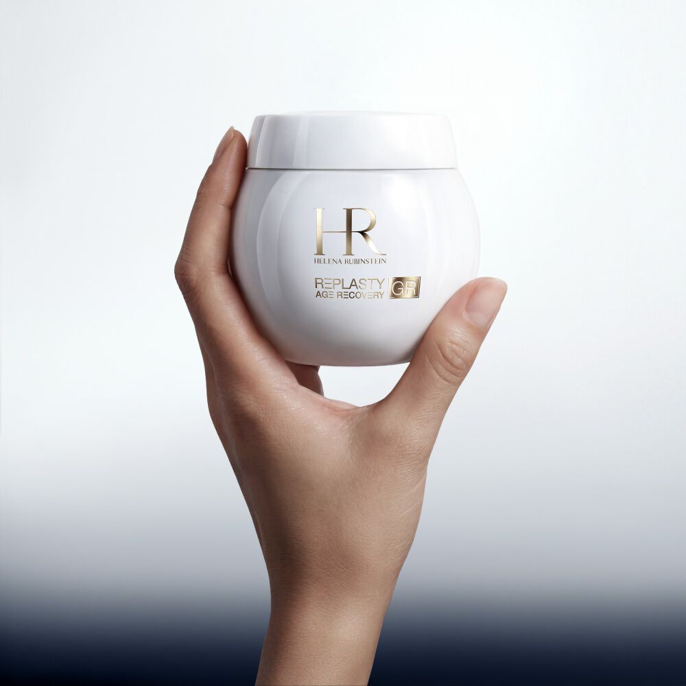 A close-up studio shot of a hand elegantly holding a glossy white round jar of Helena Rubinstein Replasty Age Recovery Day [GR] Cream. 